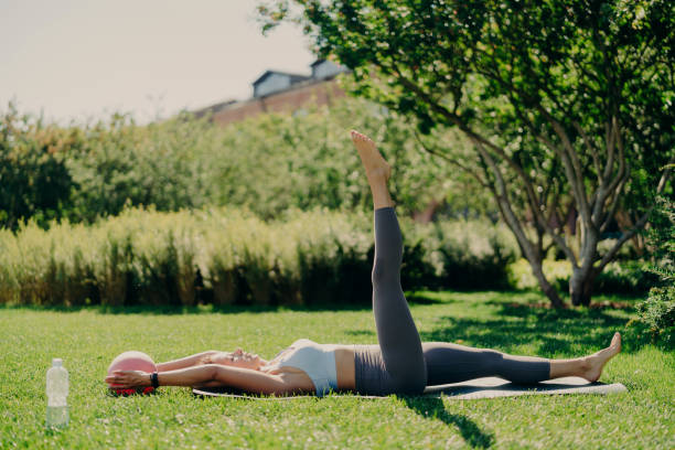 Outdoor shot of active fit woman does aerobics exercises with fitness ball lying on karemat raises leg poses on green grass with closed eyes being alone with nature enjoys tranquil atmosphere tiro al aire libre de la mujer de ajuste activo hace ejercicios de aeróbic con pelota de fitness acostado en karemat eleva las posturas de las piernas en la hierba verde con los ojos cerrados estar solo con la naturaleza disfruta de una atmósfera tranqui