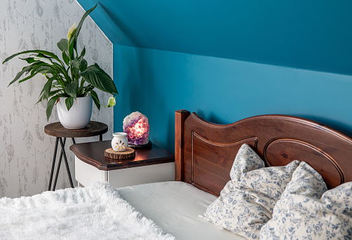 Tranquil blue color home bedroom. Air cleaning plant Spathiphyllum on flower stand, Amethyst crystal lamp illuminated and aroma lamp for relaxing aromatherapy. Natural wood furniture.