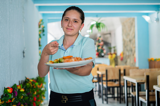 Latino woman of average age of 25 years dressed in a uniform employed by a small restaurant carries in her hand a plate from the restaurant which she shows to the camera with pride