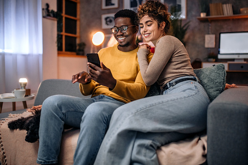 Young multiracial couple at home in the evening, resting in sofa, using phone for video call with friends