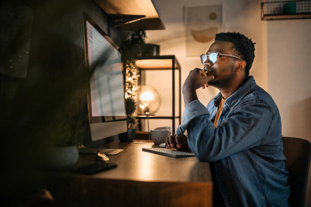 Man working late at night Young black man working late in the office, using desktop computer. working from home stock pictures, royalty-free photos & images