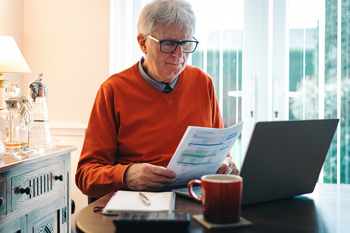 Portrait of a senior caucasian man in his 70s checking his energy bills at home. He has a worried expression while looking at the bills. Focus on the man while the interior architecture of the house is defocused.