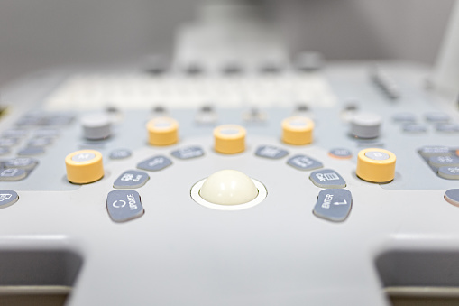 Close-up shot of medical ultrasound scanner with control bittons knobs sliders and ultrasonic transducer in background.