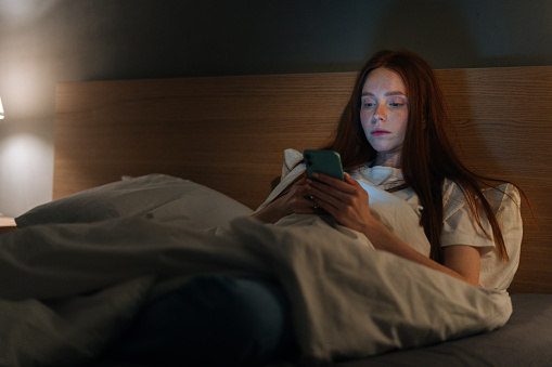 Serious insomnia young woman using smartphone, looking on screen, typing online message on social media, lying on bed late at night, bedside lamp lighting with warm yellow light.
