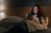 Serious insomnia young woman using smartphone, looking on screen, typing online message on social media, lying on bed late at night.