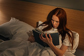 High-angle view of focused young woman reading book lying in bed at home before going to sleep. Top view of relaxed attractive redhead female reading book