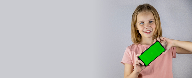 Portrait of a little blonde haired girl showing a mobile phone application in her hands with a green screen. He looks into the camera and smiles widely. Banner.