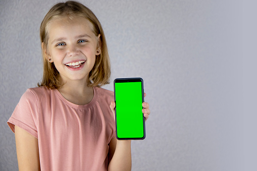 Portrait of a little blonde haired girl showing a mobile phone application in her hands with a green screen. He looks into the camera and smiles widely