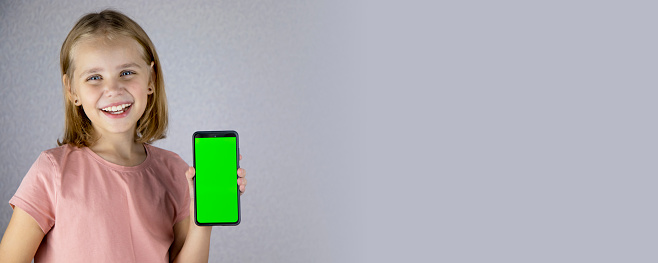 Portrait of a little blonde haired girl showing a mobile phone application in her hands with a green screen. He looks into the camera and smiles widely. Banner.