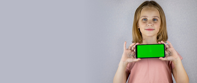 Portrait of a little blonde haired girl showing a mobile phone application in her hands with a green screen. Banner, place for text, copy space.