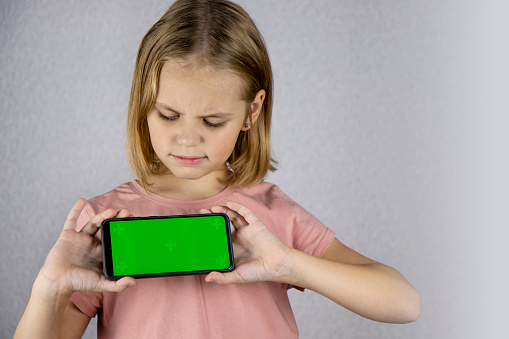 Portrait of a little blonde haired girl showing a mobile phone application in her hands with a green screen