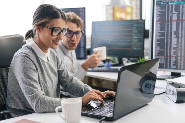 istock Young female programmer standing on her desk, coding on a laptop, having discussion with a male colleague, giving her advice 1451457118