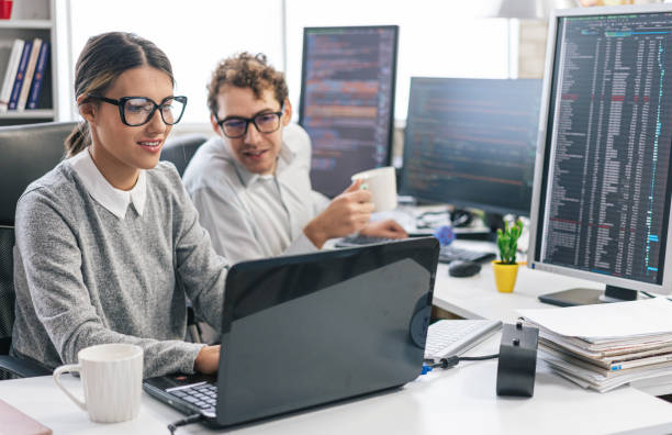 istock Young female programmer standing on her desk, coding on a laptop, having discussion with a male colleague, giving her advice 1451457123