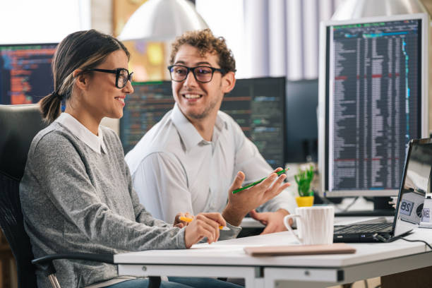 istock Young female programmer standing on her desk, coding on a laptop, having discussion with a male colleague, giving her advice 1451457166