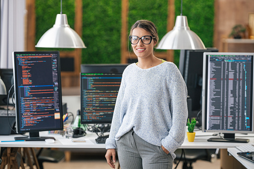 Portrait of young smiling female programmer, standing in front of her desk with computers in an IT office