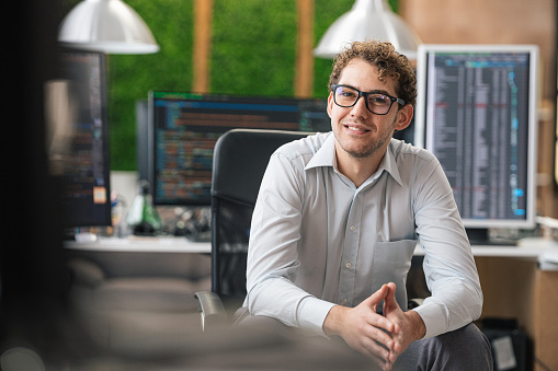 Portrait of smiling IT Programmer sitting near Desktop Computers. Male Specialist Creating Innovative Software. Engineer Developing App, Program, Video Game. Writing Code in Terminal