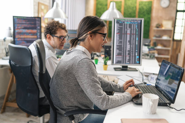 Young female programmer standing on her desk, coding on a laptop, having discussion with a male colleague, giving her advice