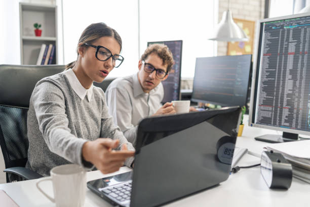 Jovem programadora em pé em sua mesa, codificando em um laptop, tendo uma discussão com um colega do sexo masculino, dando-lhe conselhos - foto de acervo