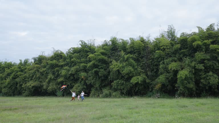 Young family with a son running together with a flying kite in a grass field by the forest in the extreme wide shot