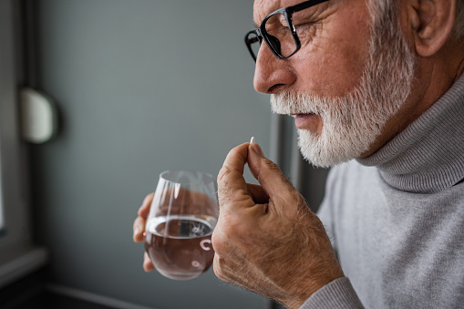 Profile view of a senior bearded man holding a glass of water and a pill