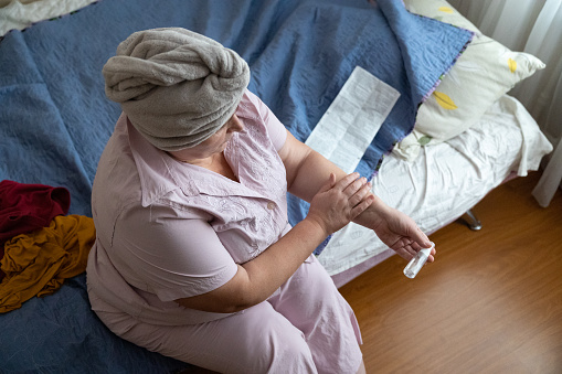 The woman rubs an estrogen spray to the inner part of the forearm. The woman is sitting on a bed in her pajamas. The woman wrapped a towel around her head to dry her hair after a shower.
