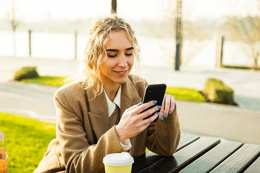 A young girl is sitting by the river on a bench and shopping via mobile phone online with shopping bags