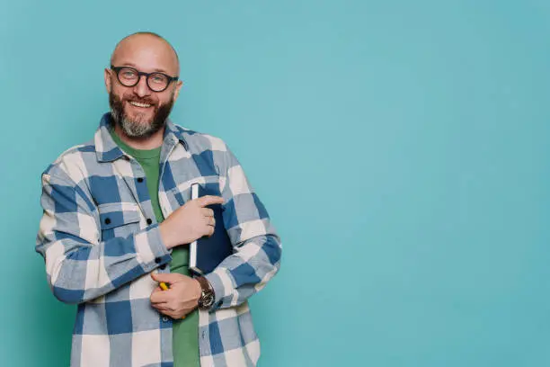 Positive Caucasian bald beardy man in plaid shirt holds book and pen looks at camera against turquoise  studio backdrop. Excited Italian male in glasses, teacher. Mockup, education, knowledge.