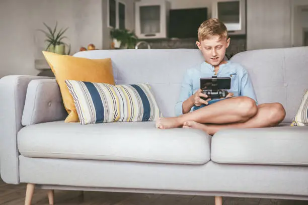 Preteen boy losted in computer game. Sits at home on coze sofa, uses gamepad with smartphone