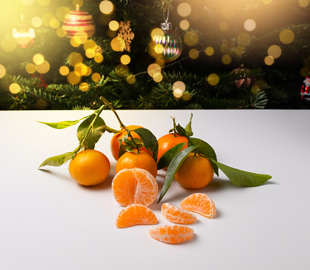 Fresh tangerines with green leaves on a white table. There is a Christmas tree with toys in the background. New Year's background. Photo