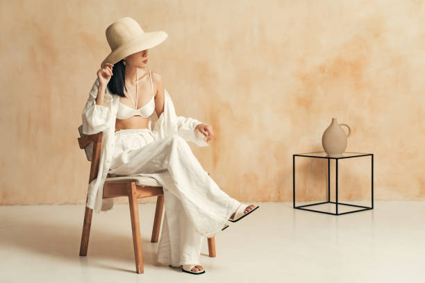 femmina asiatica irriconoscibile in grande cappello e abito da spiaggia seduta sulla sedia in arredamento minimalista - women fashion summer fashion model foto e immagini stock