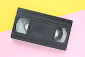 Black VHS videotape cassette on yellow and pink background. Old obsolete technology for tape recording and watching media movies