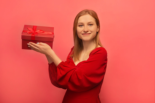 Happy smiling cheerful blonde girl in red dress holding present gift with bow and looking at camera on pink background.

Saint Valentines Day, International Women's Day or March 8 celebration concept.