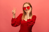 Stylish happy smiling young woman in red dress, french beret and trendy heart shaped glasses showing peace gesture v sign with two fingers and keeping hand on face on pink background.