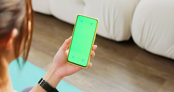 close up rear view hand fit asian woman wear sportswear and smartwatch using scrolling green screen smartphone sitting on floor at home