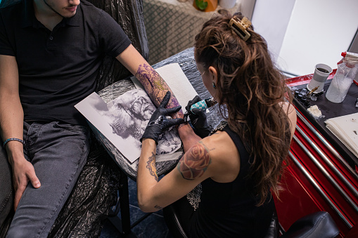 Young female tattoo artist is tattooing a man's arm in her studio.