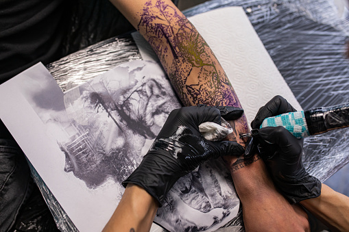 Close up photography of young man's arm which one is tattooing by young female tattoo artist.