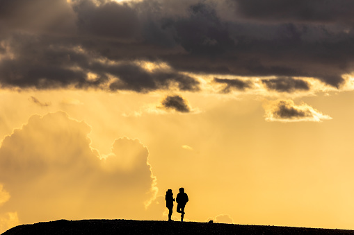 silhouette of a couple, woman and man, standing on a hill. Bright orange sunset sky with high building clouds in the background.