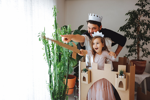 A cute little girl dressed up as a princess while playing at home. Playful childhood. Little girl and young father having fun with cardboard box.