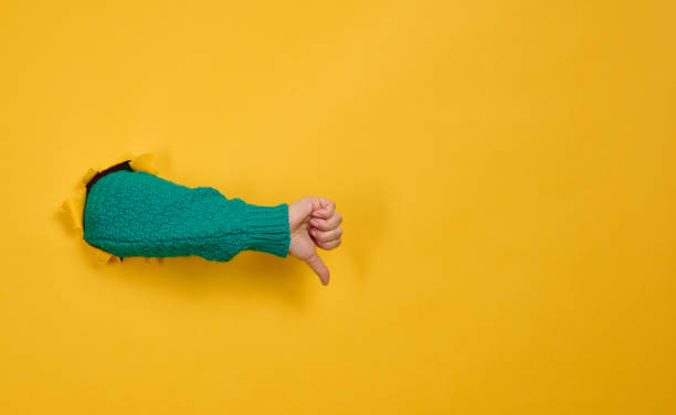 Dislike gesture break through the yellow paper. A part of the body sticks out of a hole with ragged edges Dislike gesture break through the yellow paper. A part of the body sticks out of a hole with ragged edges. Copy space thumb down stock pictures, royalty-free photos & images
