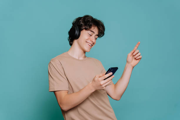 handsome spanish young guy listening music uses headphones holds phone eyes closed against blue studio backdrop. joyful schoolboy dancing smiles wide. happy teenager loves music. mockup, youth. - alleen één tienerjongen stockfoto's en -beelden