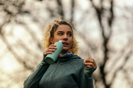 Young woman relaxing and drinking water after jogging in nature on an autumn day. She has a training in public park in urban area