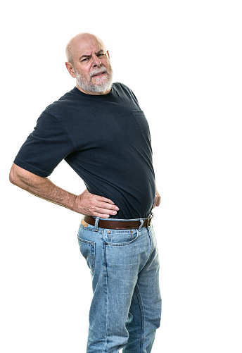 Muscular senior adult man with back aches and pains is grimacing at the camera as he twists his upper body to stretch his back muscles while pushing and massaging around his hips and lower back with his hands as he tries to get more comfortable. White background.