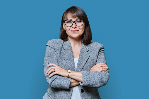 Business positive middle age woman with arms crossed on blue studio background. Confident woman with smile in glasses jacket looking at camera. Business success management leadership staff 40s people