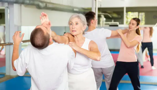 Senior European woman learning chin strike move on man during self-defense training. Othe woman training with young man in background.