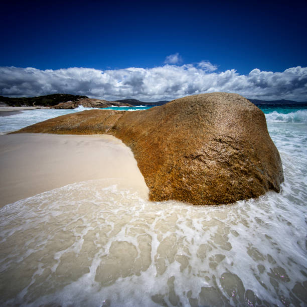 Waves wash around a smoothed granite boulder on white beach sand with blue sky