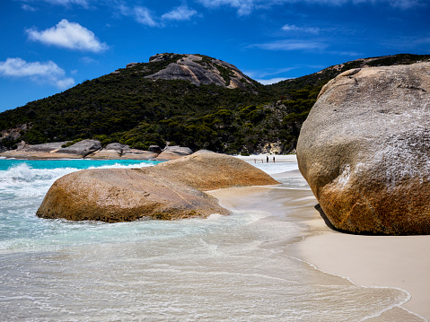 Waves wash around smoothed granite boulders on white beach sand with blue sky and bush covered mountain behind