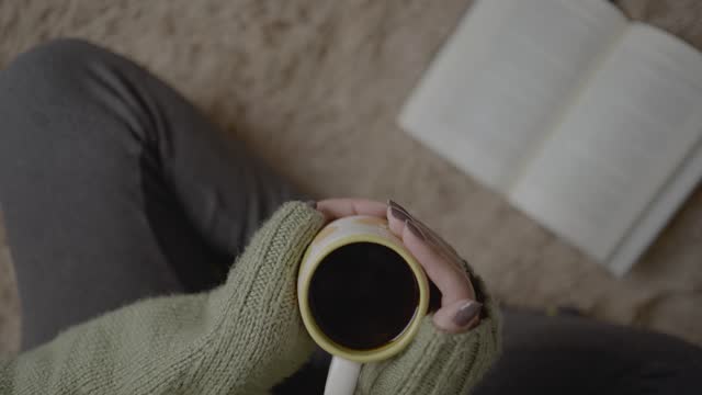 First person angle view of woman wearing wool sweater holding cup of coffee while reading a book on the living room carpet floor