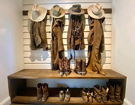 Horizontal still life of country style cowboy boots with horse riding spurs, riding jackets and hats with bridle on wood wall hook and bench  on rural farm horse property Bangalow NSW Australia