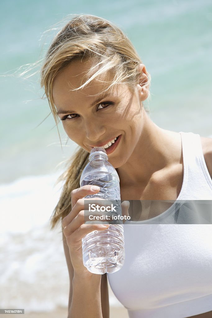 Smiling woman drinking from a bottle of water on the beach Smiling young caucasian female drinking bottled water with ocean water behind her. One Woman Only Stock Photo