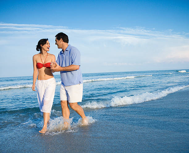 Couple walking in waves. Caucasian young adult man and woman holding hands laughing and wading in ocean. bald head island stock pictures, royalty-free photos & images